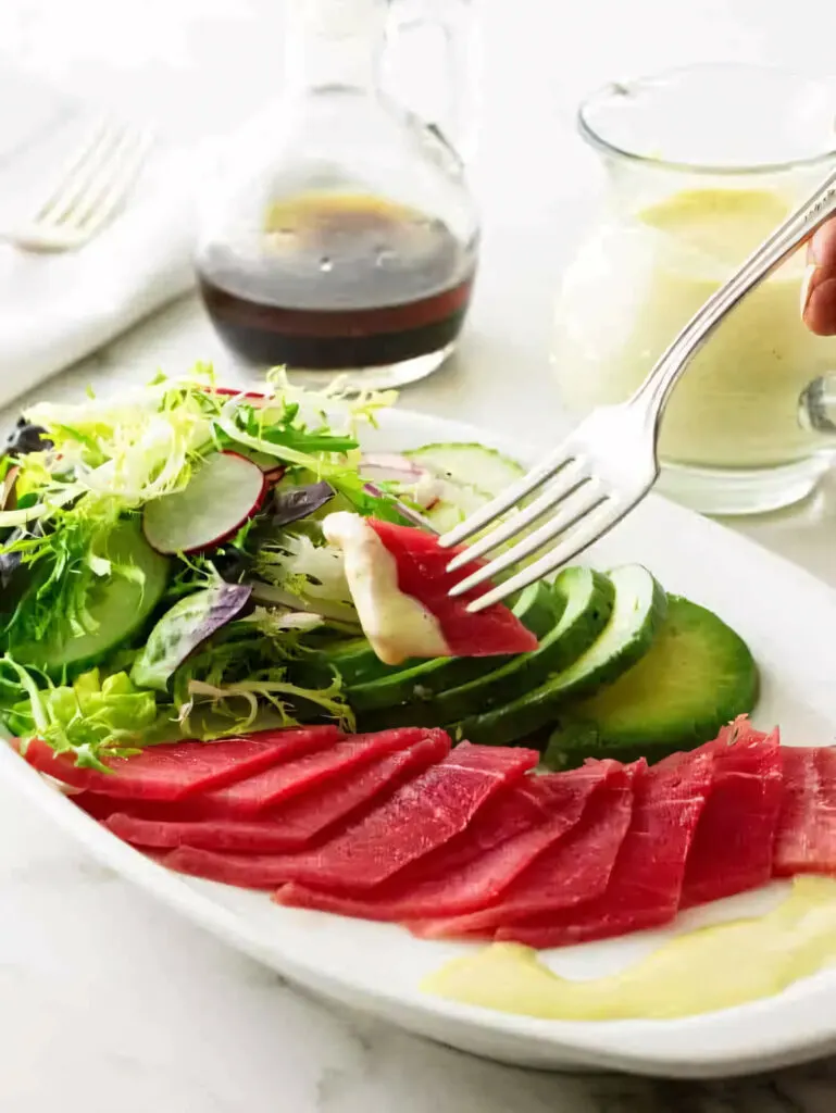 A fork grabbing a bite of tuna sashimi and dipping it in wasabi dressing.