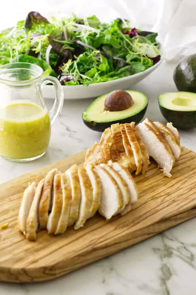 Sliced chicken breast with mixed green salad and avocado in the background.