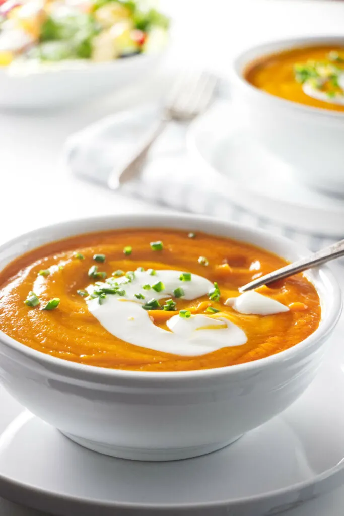 A bowl of carrot soup with a salad in the background.