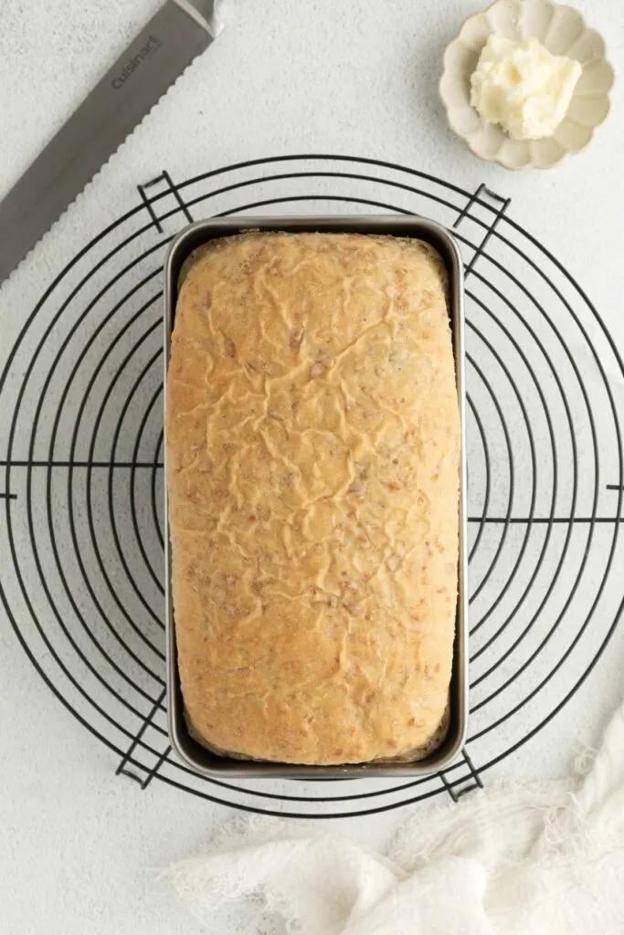 A loaf of bread in a pan cooling on a rack next to butter.