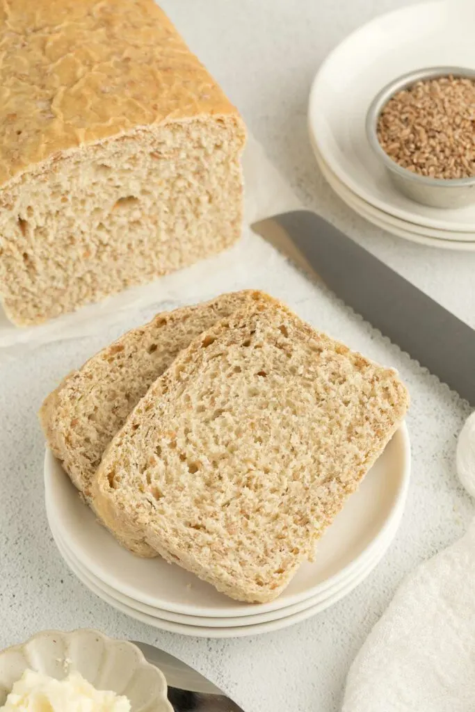 A couple of slices of bread next to a cup of cracked wheat and a knife.