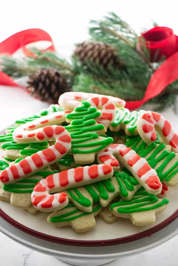 A plate of Christmas cut out cookies in front of pine cones and red ribbons.