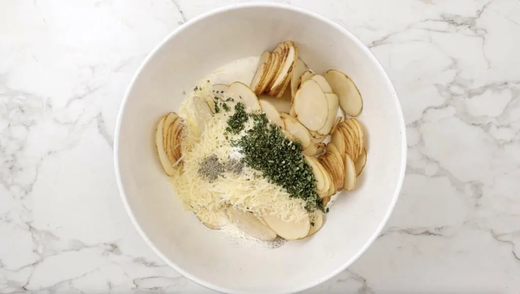 Thinly sliced potatoes in a bowl with cream, parmesan, rosemary, salt, and pepper.