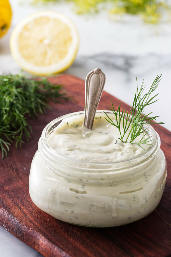 A jar filled with homemade lemon dill mayo and a sliced lemon in the background.