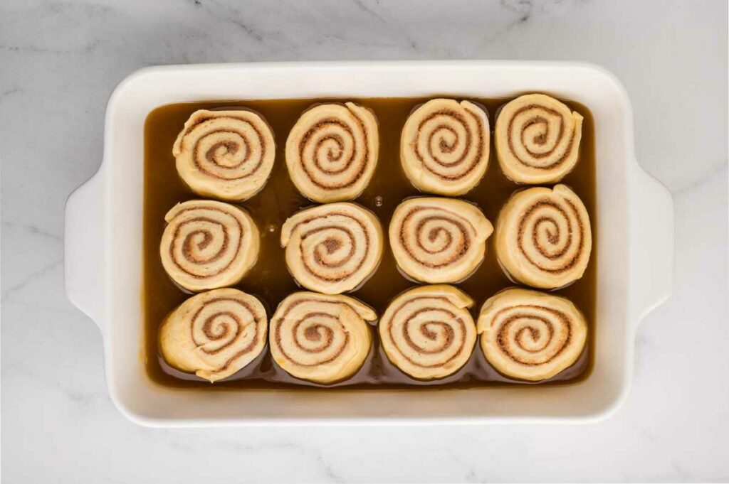 Unproofed cinnamon rolls arranged in a baking pan on top of a layer of sticky caramel sauce, ready to rise and bake.