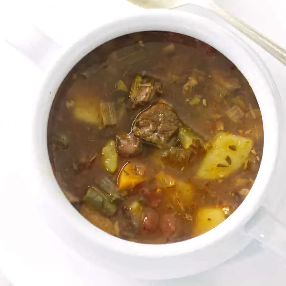 A bowl of beef and vegetable soup next to a spoon.