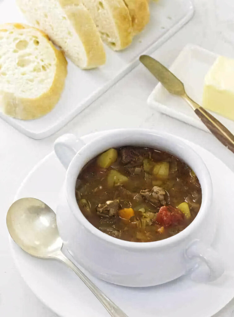 A bowl of beef and vegetable soup next to a loaf of bread.