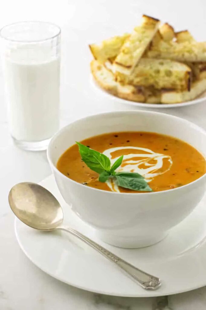 A bowl of roasted tomato soup in front of a glass of milk and some bread.