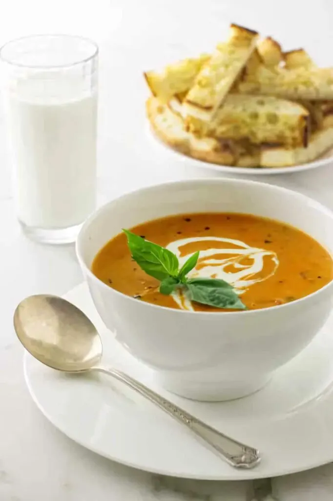 A bowl of roasted tomato soup in front of a glass of milk and some bread.