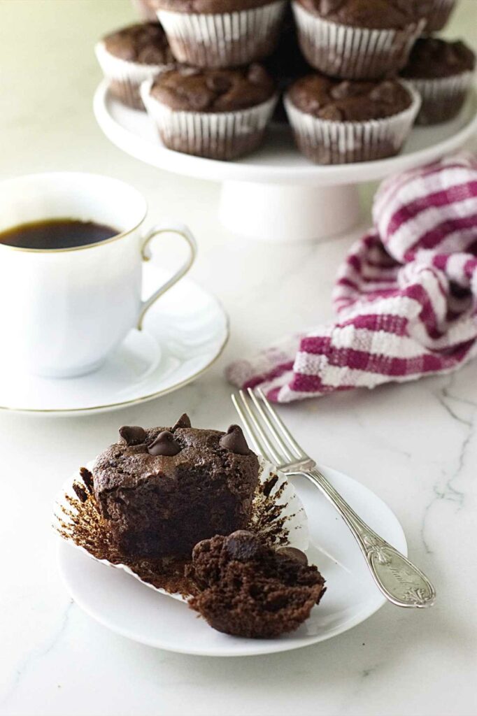 An einkorn banana chocolate muffin on a plate with a fork and a cup of coffee in the background.