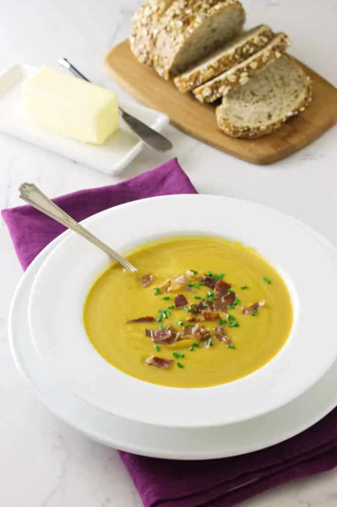 A bowl of butternut prosciutto soup next to a loaf of sliced bread.