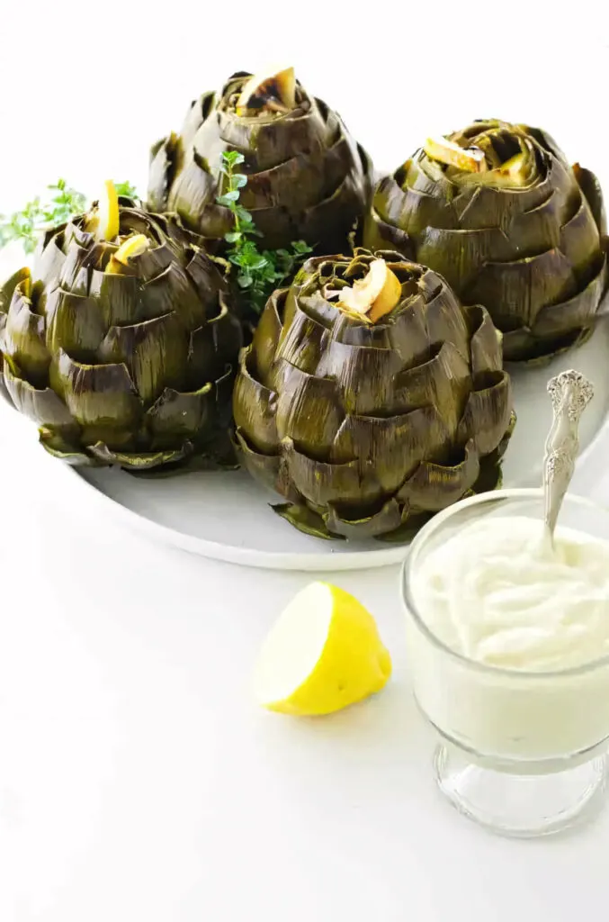 Four oven baked artichokes on a serving plate in front of a dish of aioli.