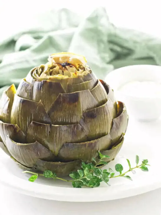 A roasted artichoke on a plate with a sprig of thyme.