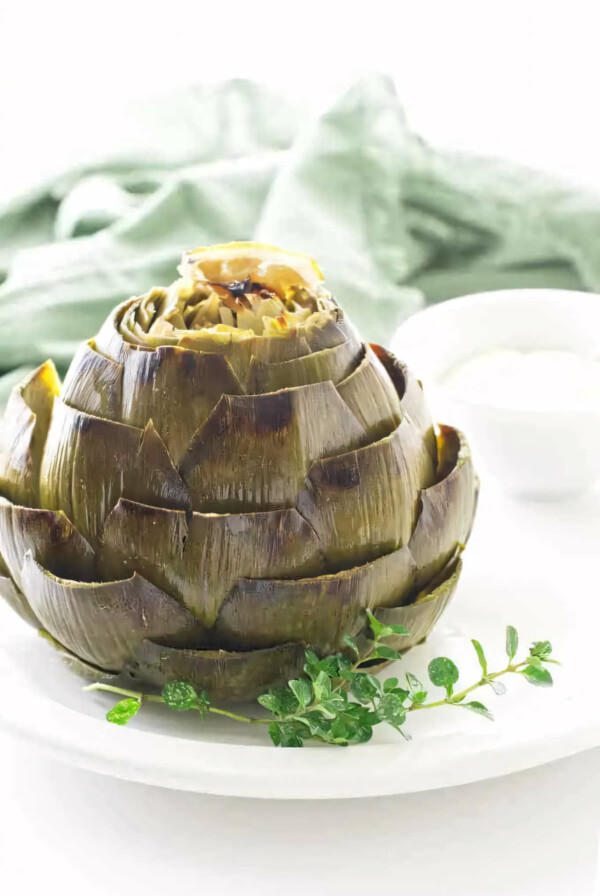 A roasted artichoke on a plate with a sprig of thyme.