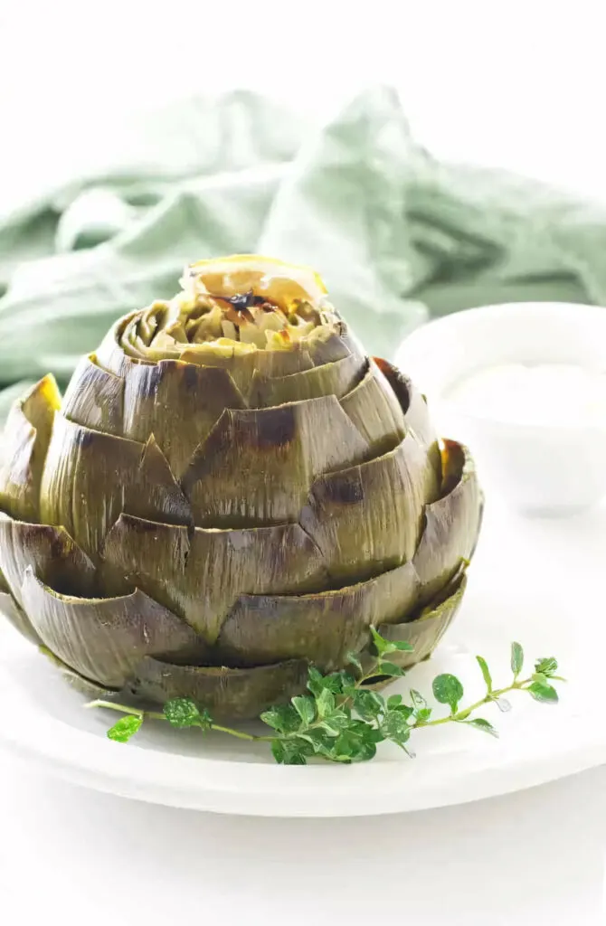 A roasted artichoke on a plate with a sprig of thyme.