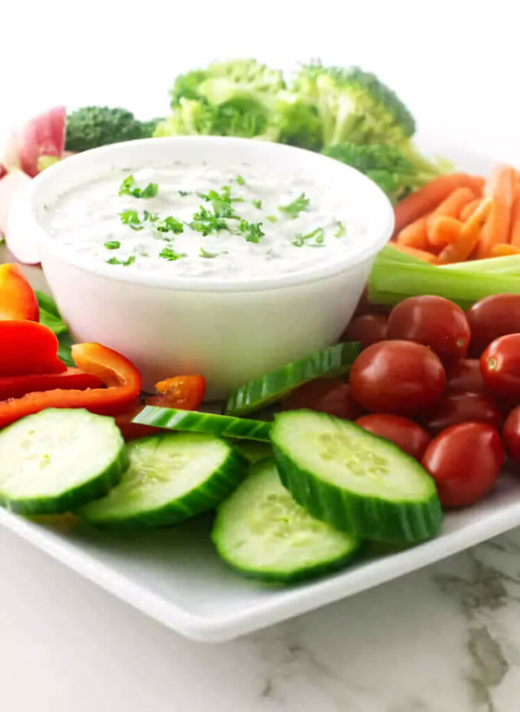 A bowl of Greek yogurt dip on a platter with carrots, tomatoes, celery sticks, and cucumber.