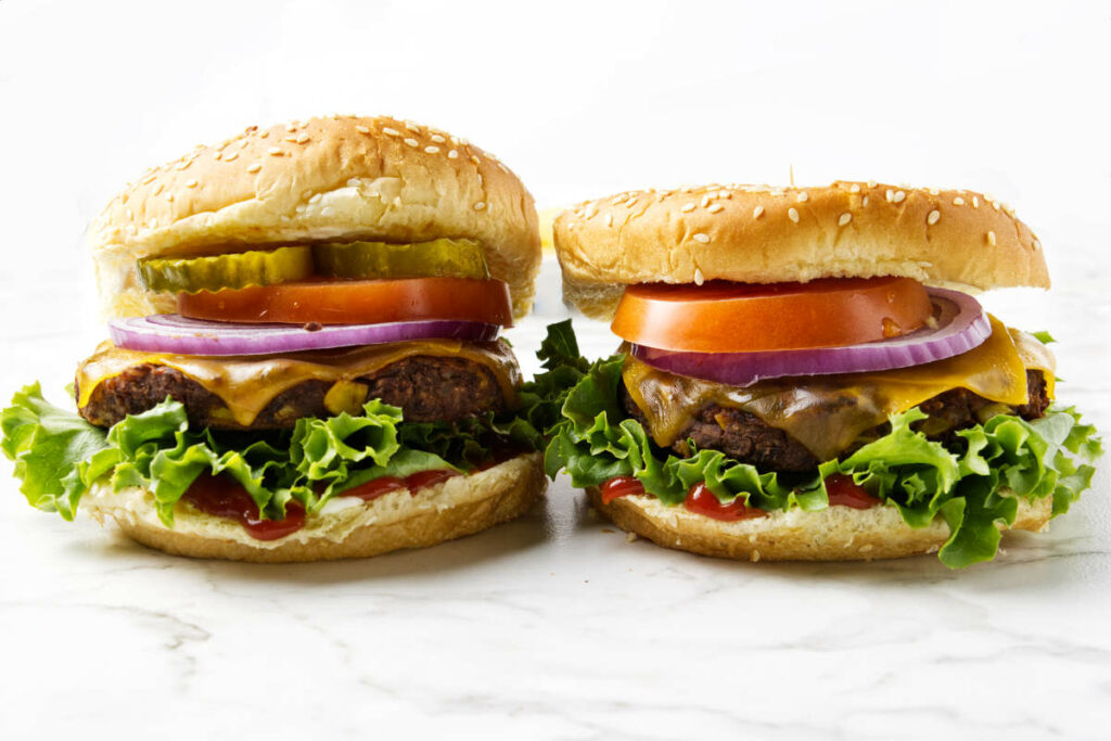 Two burgers next to each other with lettuce, onion, and tomatoes.