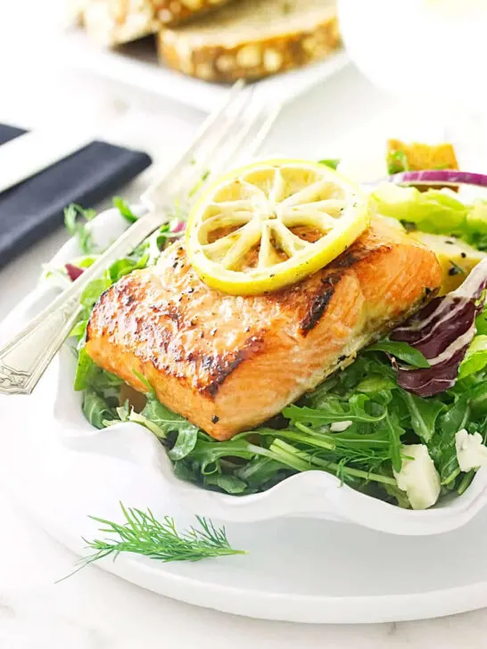 A salad plate filled with salmon and salad greens with bread in the background.