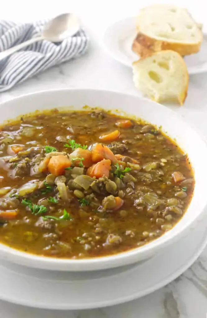 A bowl of lentil soup on a plate with a serving of bread in the background.