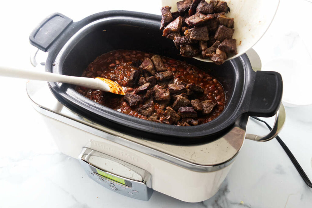 Adding cubes of browned beef to a slow cooker filled with chili.