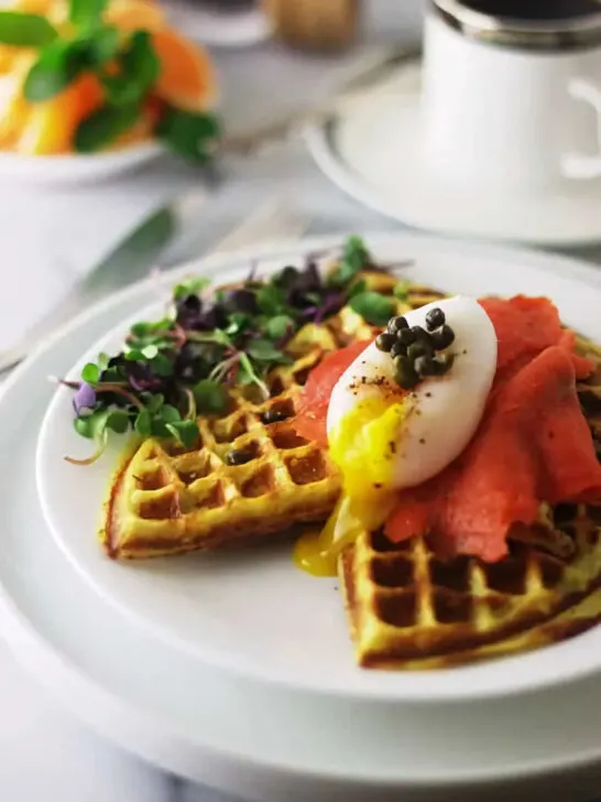 A sweet potato waffle topped with smoked salmon and an egg in front of a cup of coffee.