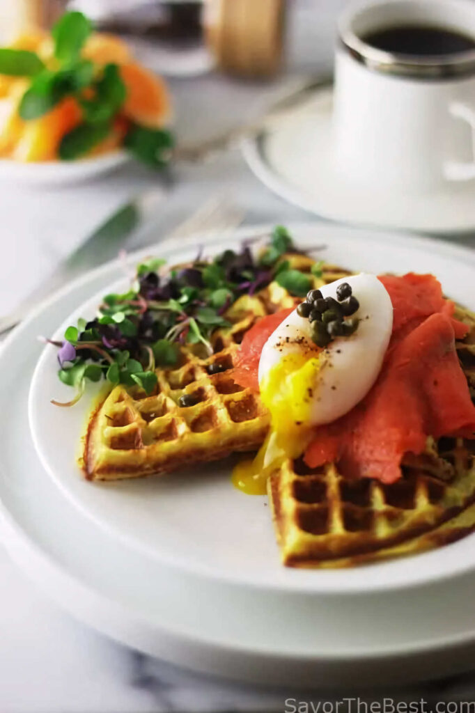 A sweet potato waffle topped with smoked salmon and an egg in front of a cup of coffee.