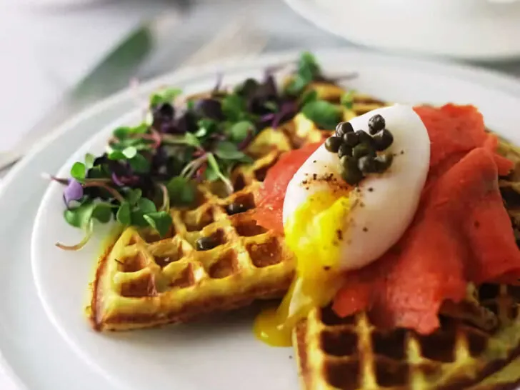 A sweet potato waffle topped with smoked salmon and an egg in front of a cup of coffee.