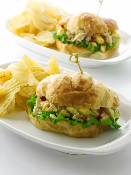 Two plates with a chicken croissant sandwich next to potato chips.