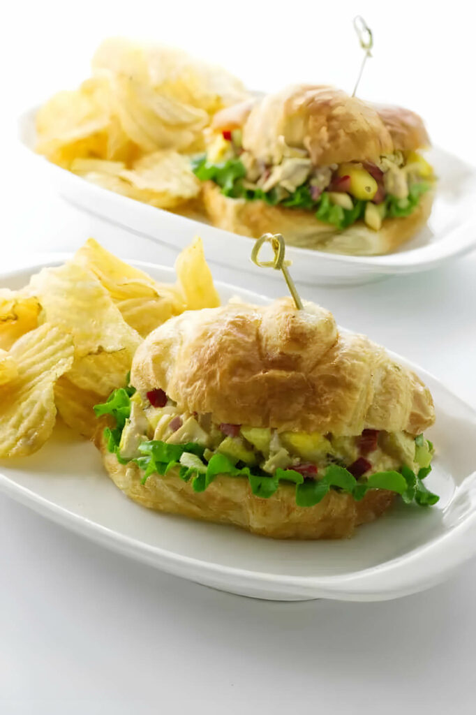 Two plates with a chicken croissant sandwich next to potato chips.