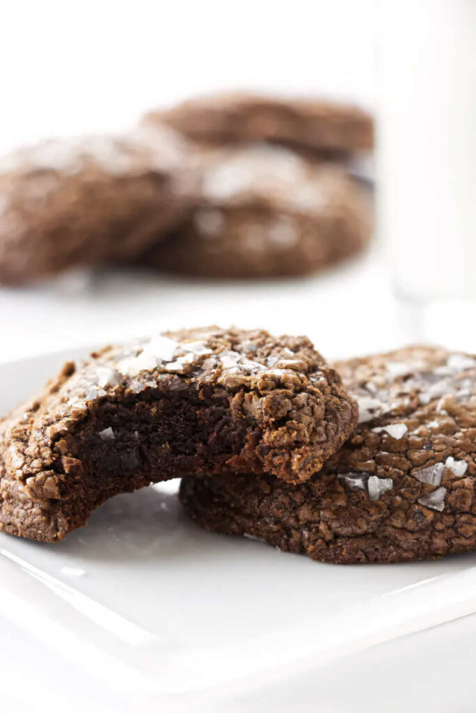 A partially eaten chocolate fudge cookie on a plate with a whole cookie.