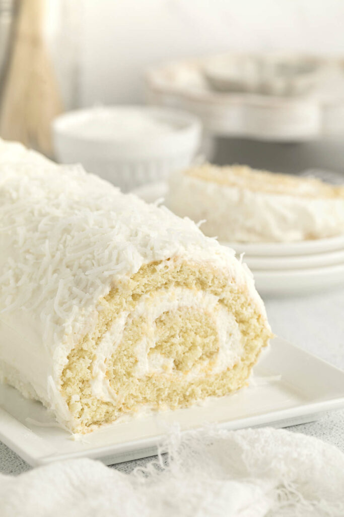 A coconut roll on a platter with a slice of cake in the background.