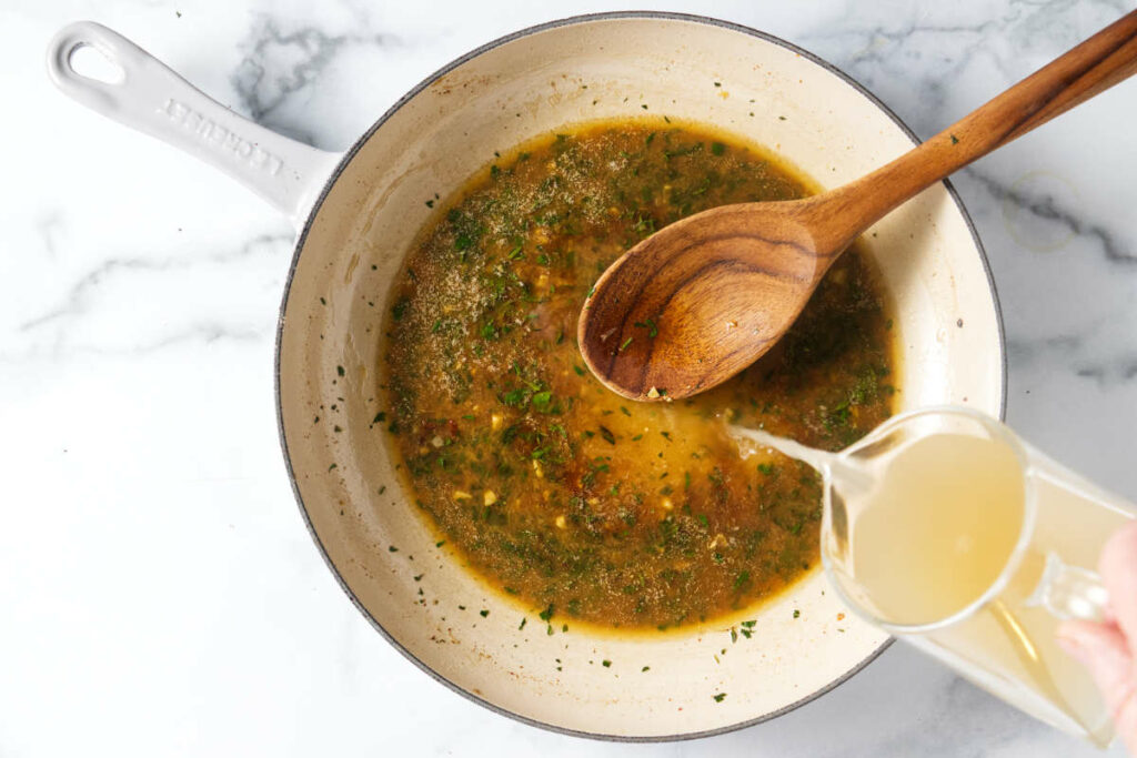 Adding chicken broth to the skillet with herbs.