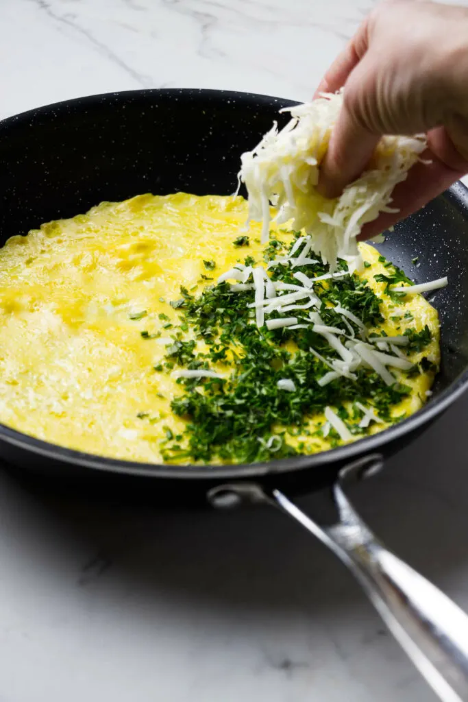 Adding herb and grated cheese on half of the skillet with an omelet.