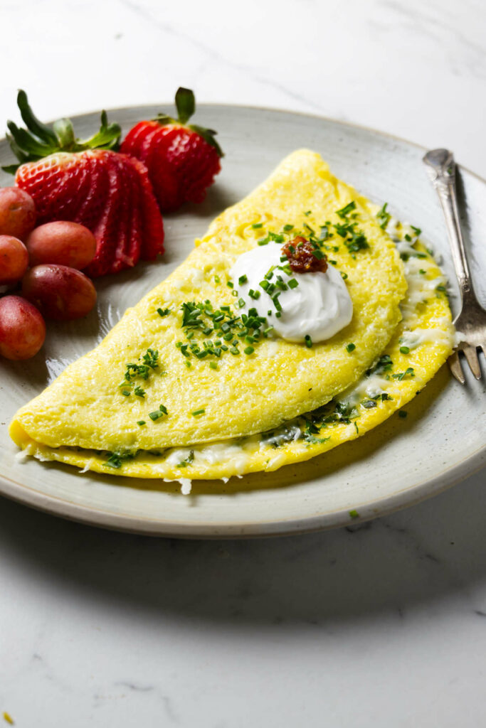 A three egg omelet on a plate with fruit then topped with fresh herbs.