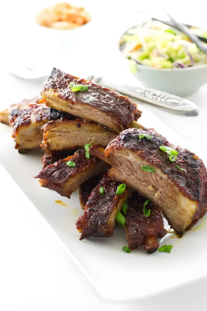 A platter filled with slices of Asian sticky pork ribs with kimchi in the background.