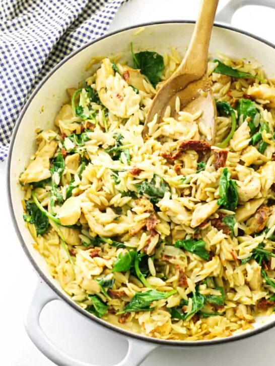 A skillet with chicken orzo and kale along with a wooden spoon.