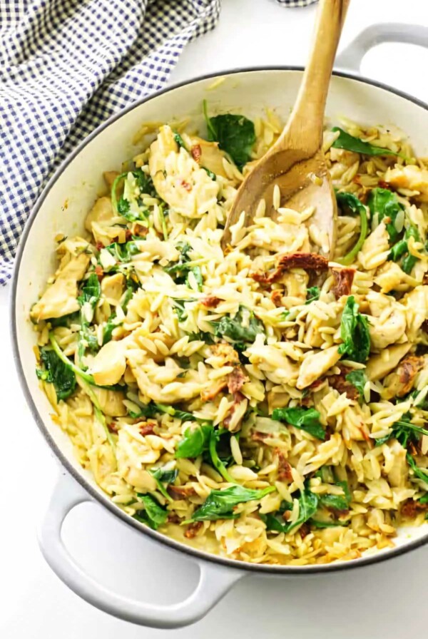 A skillet with chicken orzo and kale along with a wooden spoon.