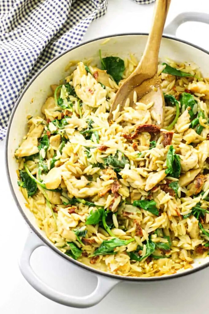 A skillet with chicken orzo and kale along with a wooden spoon.