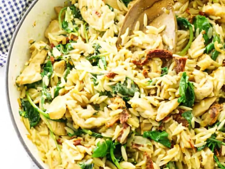 A skillet with chicken orzo and kale along with a wooden spoon.