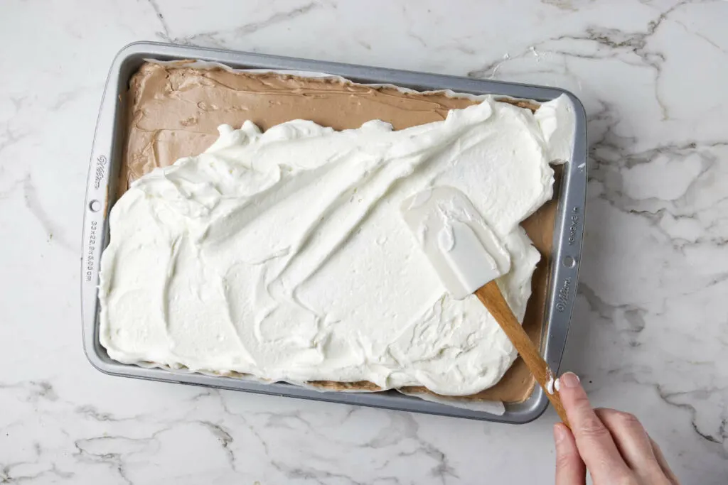 Spreading whipped cream on top of a chocolate caramel layered cheesecake bar.