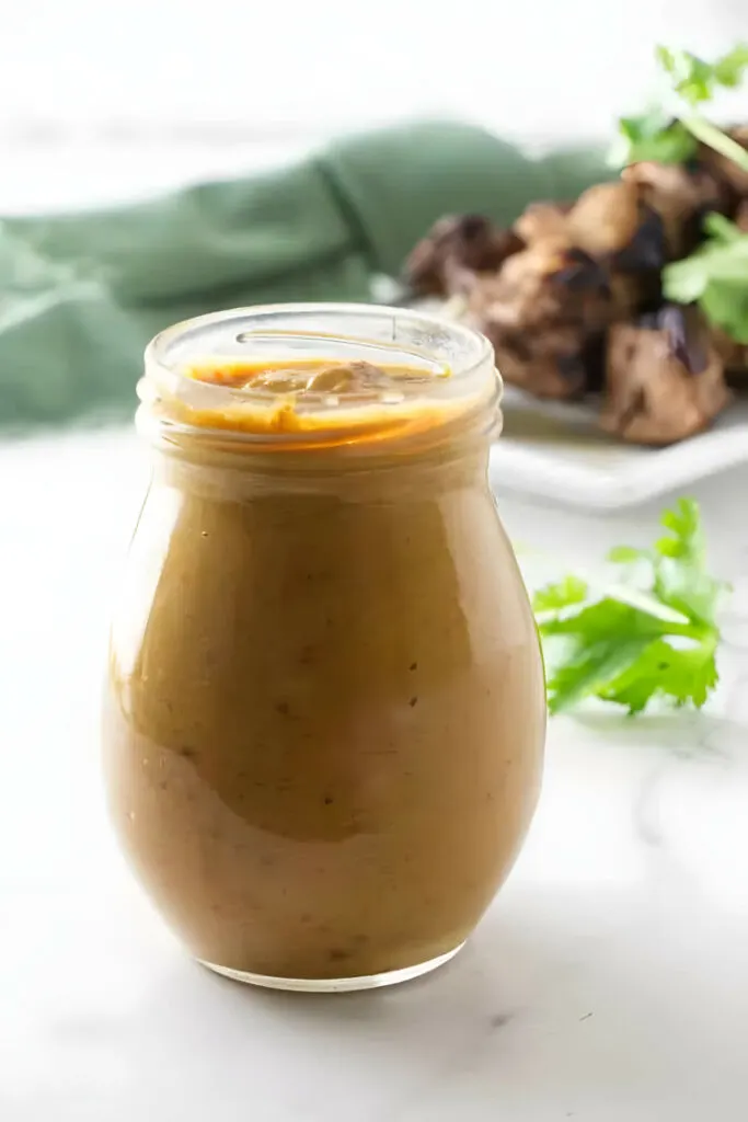 A jar filled with Thai peanut sauce and chicken kabobs in the background.