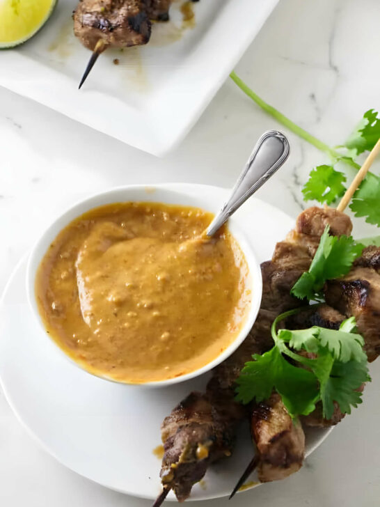 A small dish of spicy peanut butter sauce next to chicken and herbs.