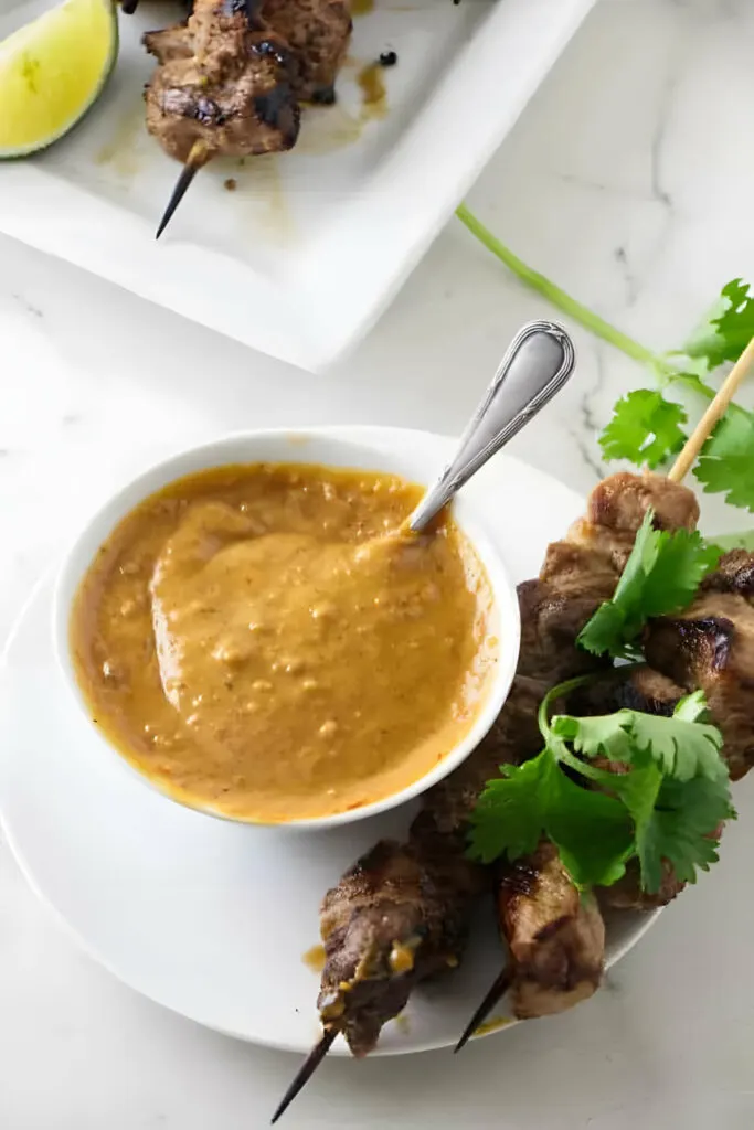 A small dish of spicy peanut butter sauce next to chicken and herbs.