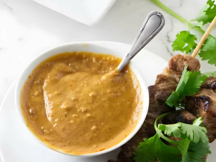 A small dish of spicy peanut butter sauce next to chicken and herbs.