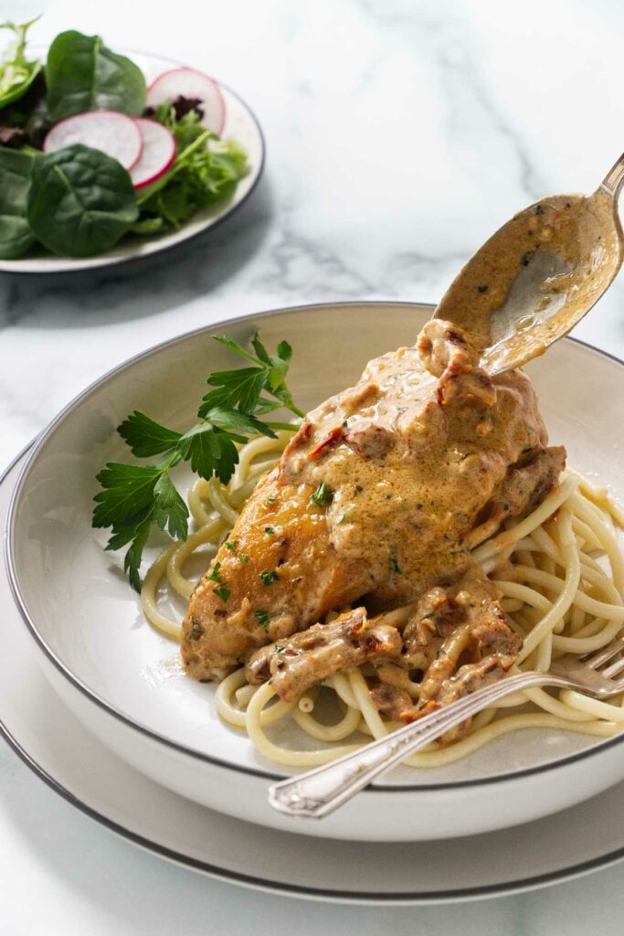 Spooning creamy tomato sauce over a serving of chicken with a green salad in the background.