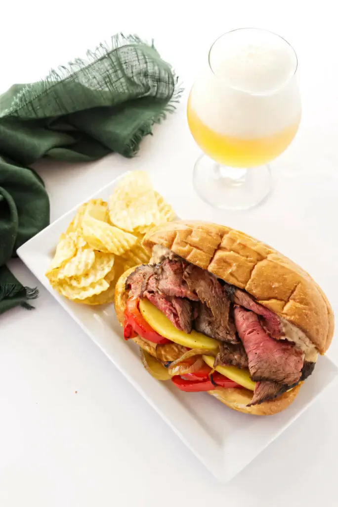 A tri tip sandwich on a hoagie bun next to potato chips and a drink.