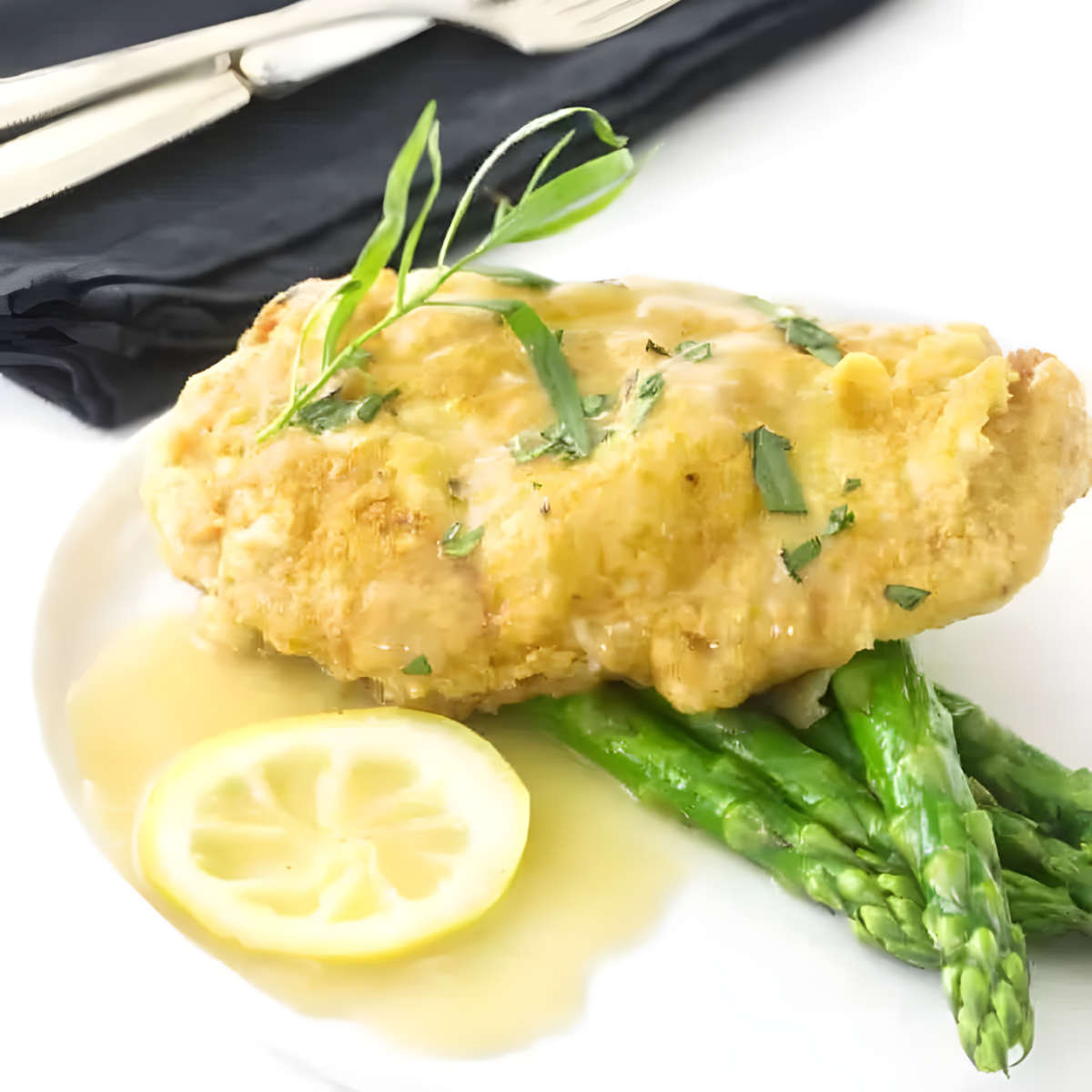 A pan fried chicken breast on a plate with lemon sauce and asparagus.