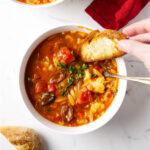 Chicken cacciatore orzo soup in a bowl next to a slice of bread.