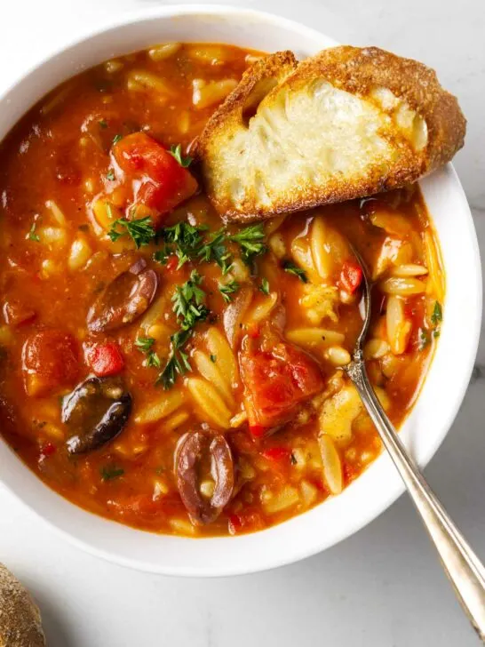 A spoon in a bowl of chicken cacciatore orzo soup along with a slice of French bread.