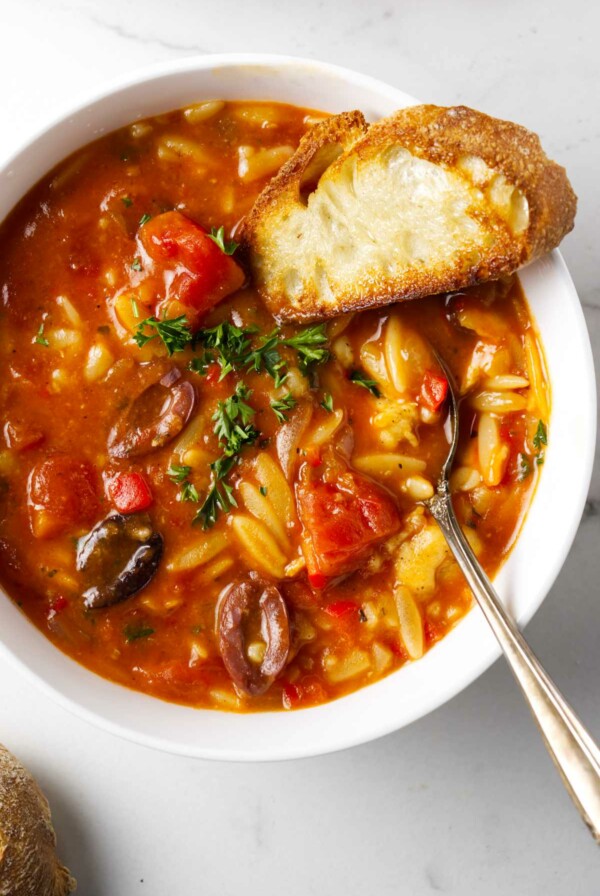 A spoon in a bowl of chicken cacciatore orzo soup along with a slice of French bread.